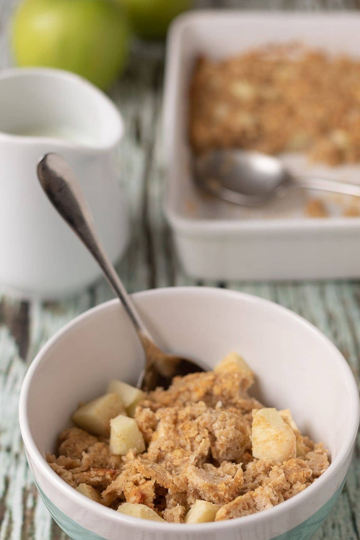 Bowl of cooked baked apple and cinnamon oats served with spoon in. Rest of the serving dish and a jug of milk and an apple in the background.