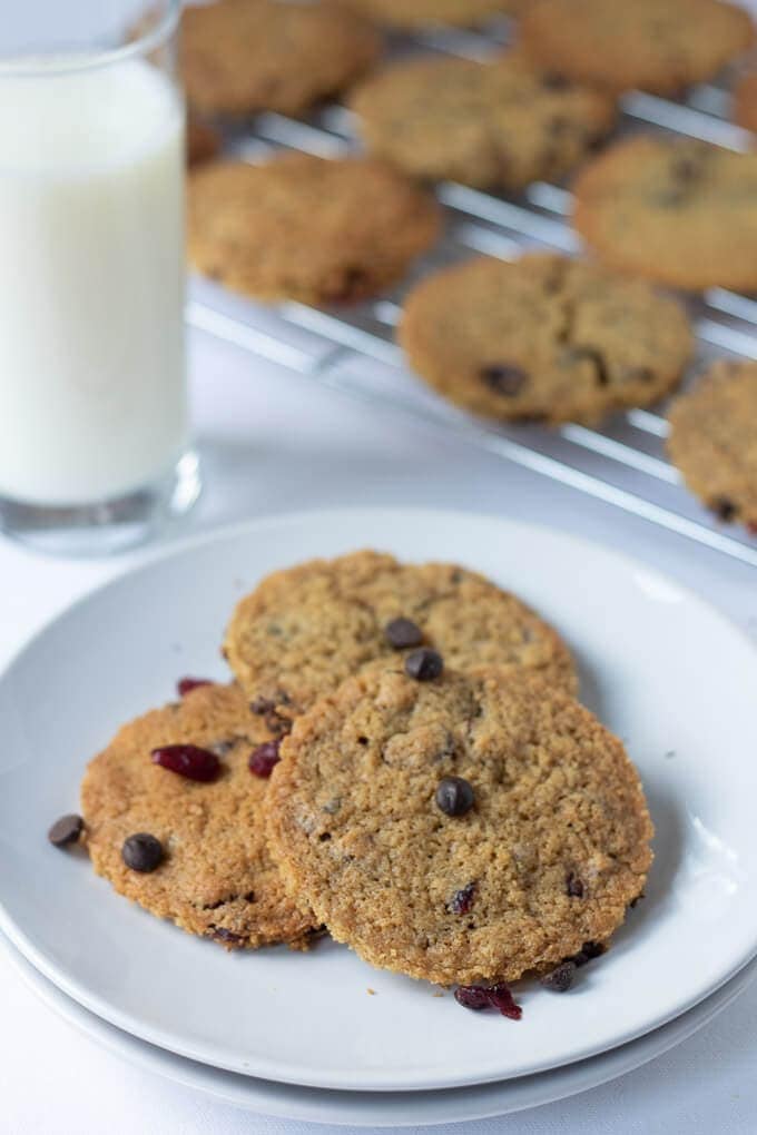 Plate of 3 delicious dark chocolate chip cranberry cookies with a glass of milk and rack of cookies behind.