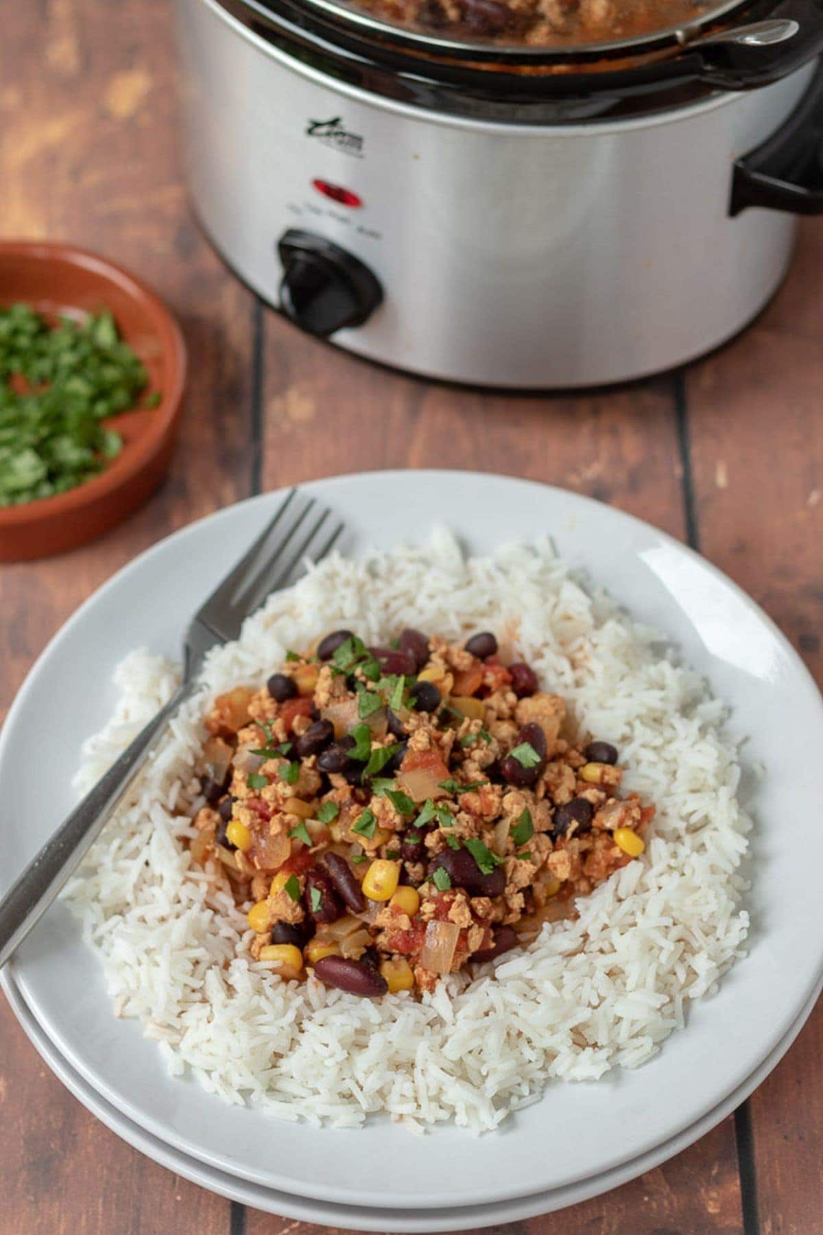 Healthy slow cooker turkey chilli on a plate served on rice and garnished with coriander. Slow cooker in the background with a dish of chopped coriander in between.