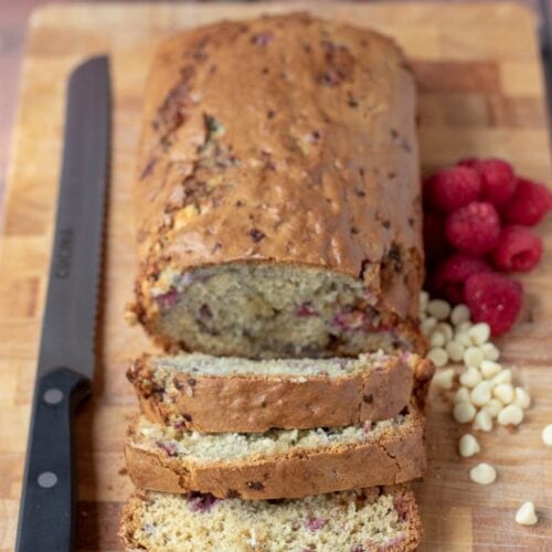 White chocolate and raspberry loaf cake on a bread board facing forward with three slices cut off. Carving knife to the left hand side.