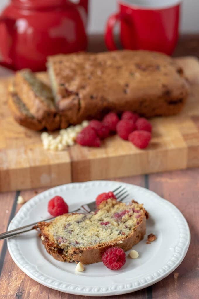 Close up of a slice of white chocolate and raspberry loaf cake on a plate with the loaf on a bread board in the background with 2 slices cut off.