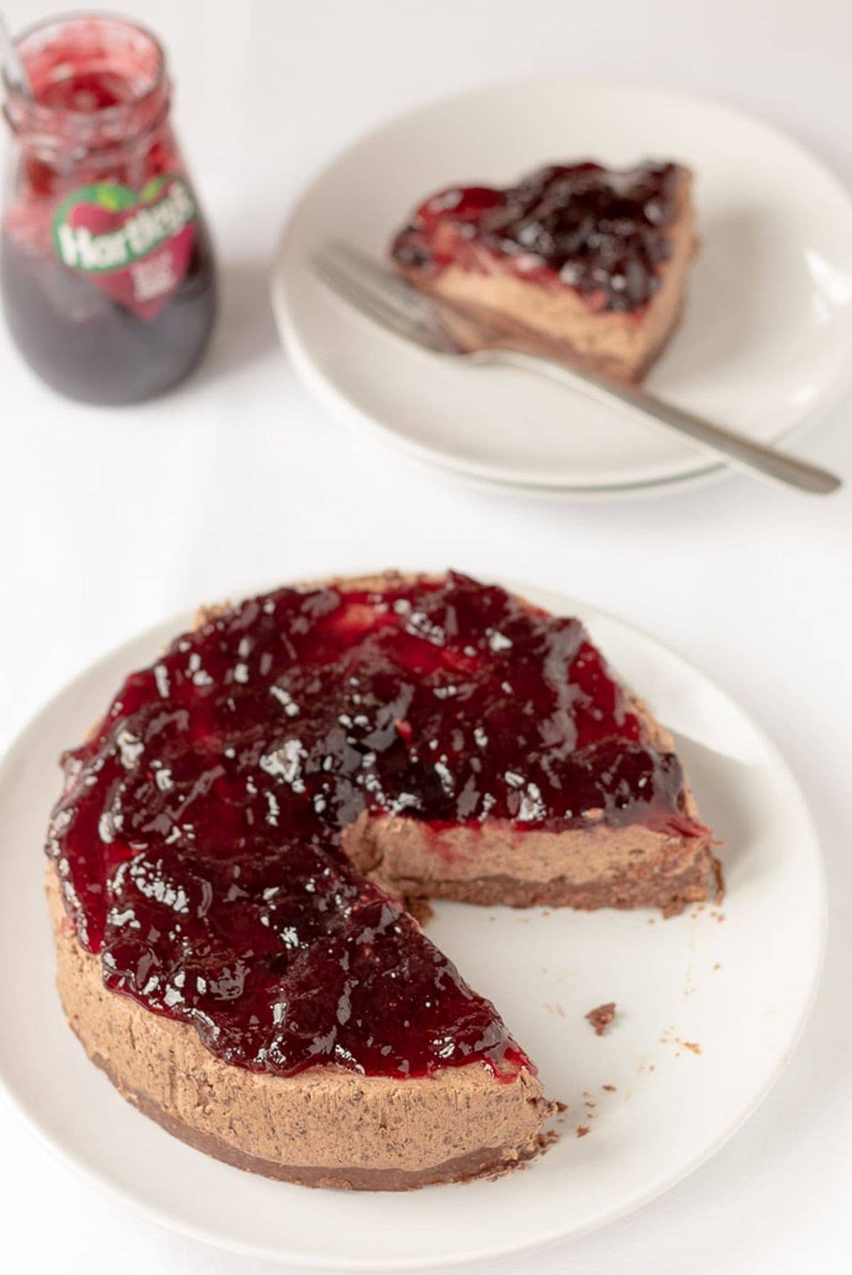 Delicious no-bake black cherry chocolate cheesecake on a plate with a slice taken out of it and placed on a plate to the rear with a serving fork on. A jar of jam to the left side.