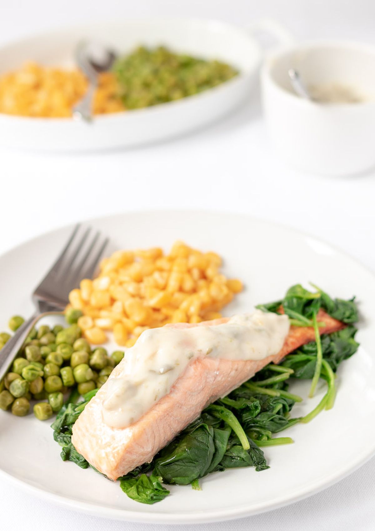 Healthy grilled salmon served on a bed of wilted spinach with a portion of sweetcorn and peas on a plate.