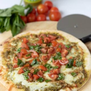 A delicious pesto pizza cooked sitting on a chopping board, ready to carve into slices and eat with cherry tomatoes and basil in the background for decoration.
