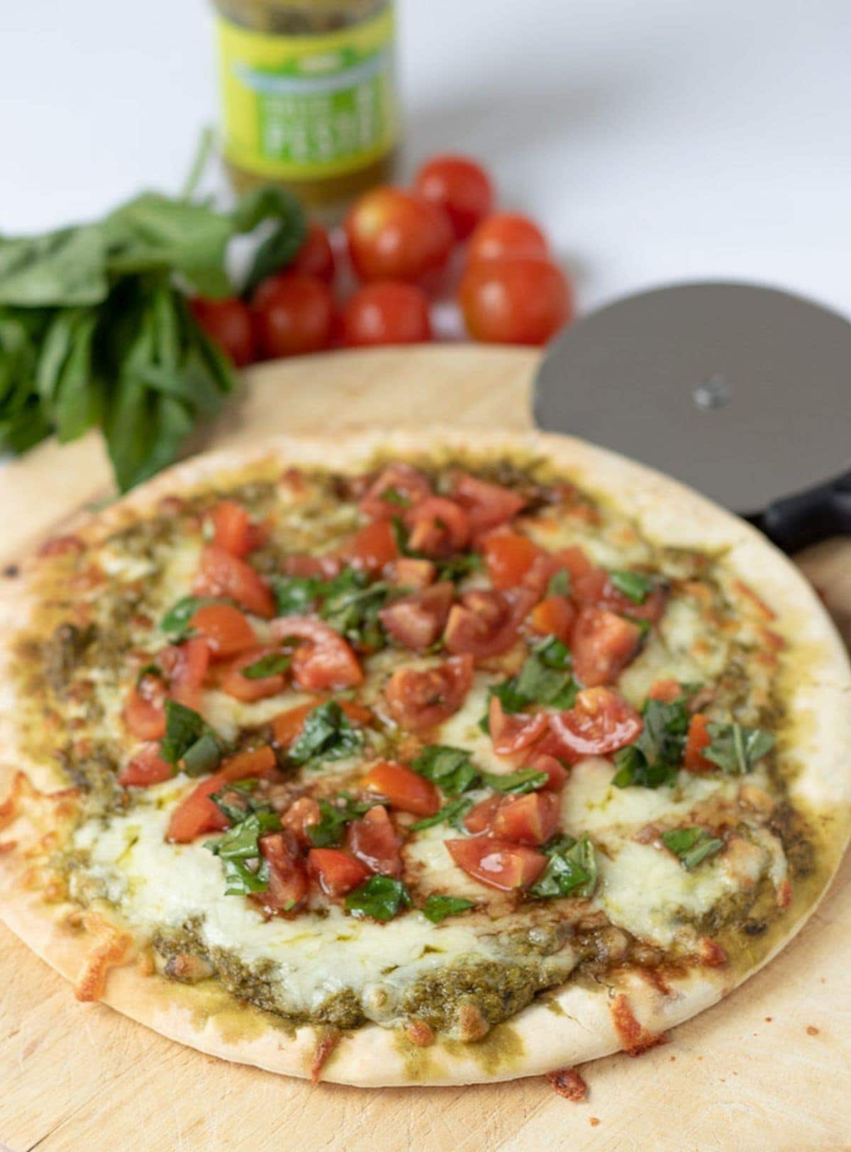 A delicious pesto pizza cooked sitting on a chopping board, ready to carve into slices and eat with cherry tomatoes and basil in the background for decoration.