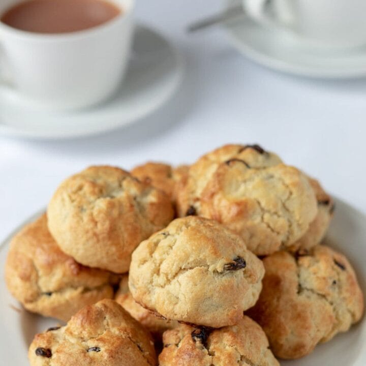 Rock cakes in a pile on a plate with cups of freshly pured tea in the background.