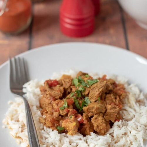 Delicious slow cooker beef curry on a plate served on a bed of rice garnished with coriander and a fork beside. SAlt and pepper cellars, a bowl of rice and spice jar in the background.
