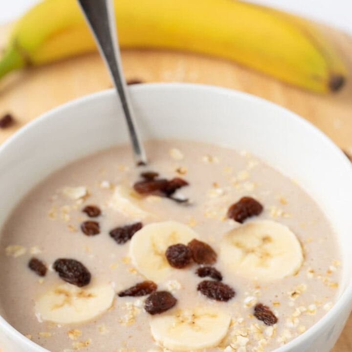 A bowl of delicious banana cinnamon overnight oats topped with slices of banana and a spoon in, ready to eat with a banana in the background as decoration.