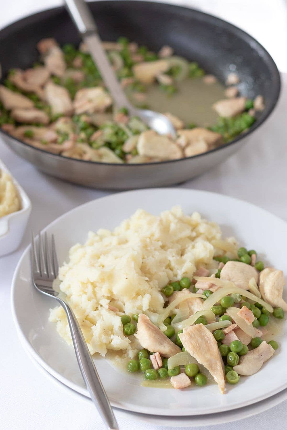 Simple chicken with peas and bacon served on a plate with creamy mashed potato with the cooking pan in the background.
