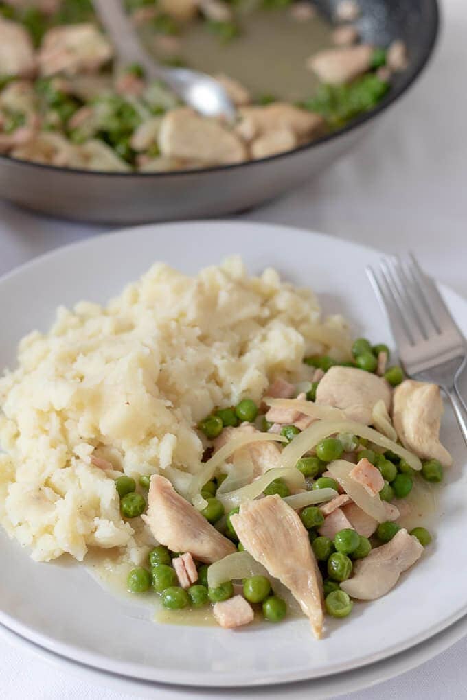 Close up view of a delicious plate of simple chicken with peas and bacon served with creamy mashed potato and served with the cooking pan in the background.