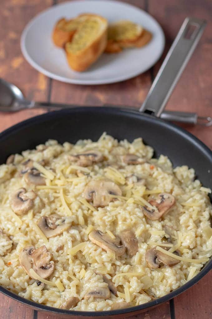A pan of the cooked mushroom risotto sits in front of a plate with two slices of garlic bread on.