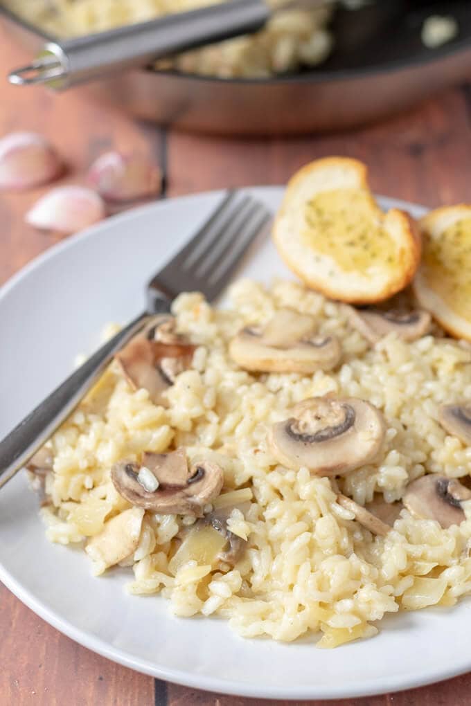 A plate of cooked and served mushroom risotto with a folk and 2 slices of garlic bread on it ready to eat.