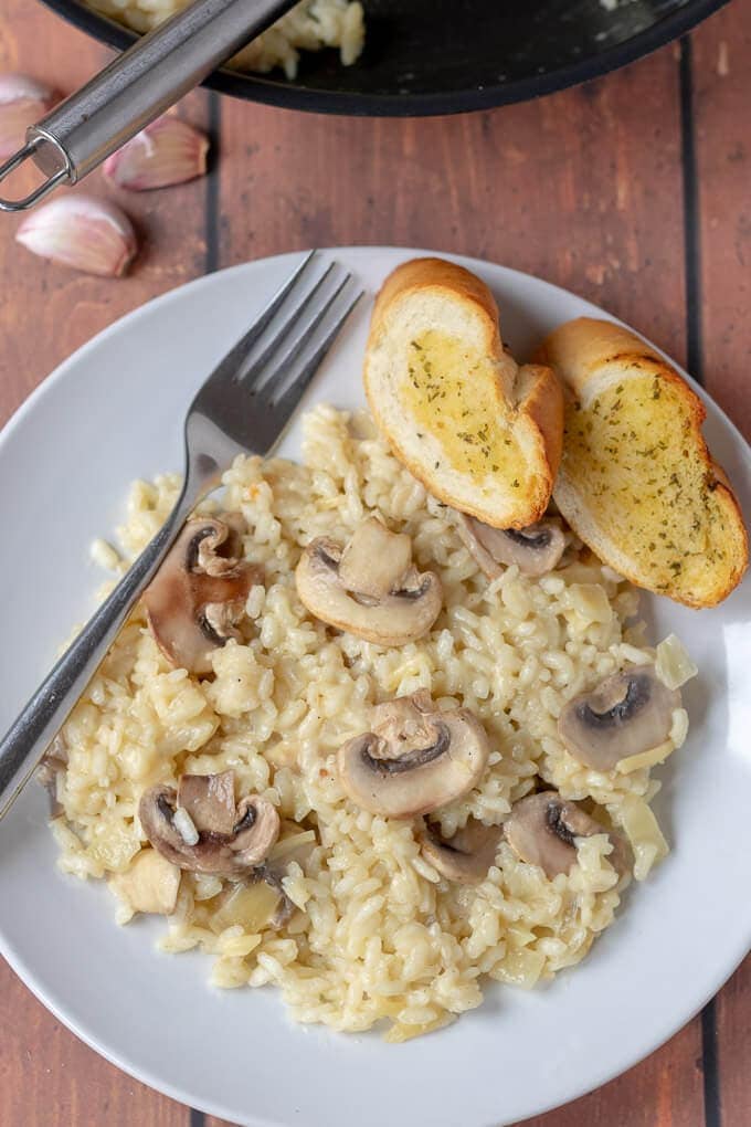 Birds eye view of a plate of cooked and served 30-minute mushroom risotto with a fork on the side ready to eat. Served with 2 slices of garlic bread.