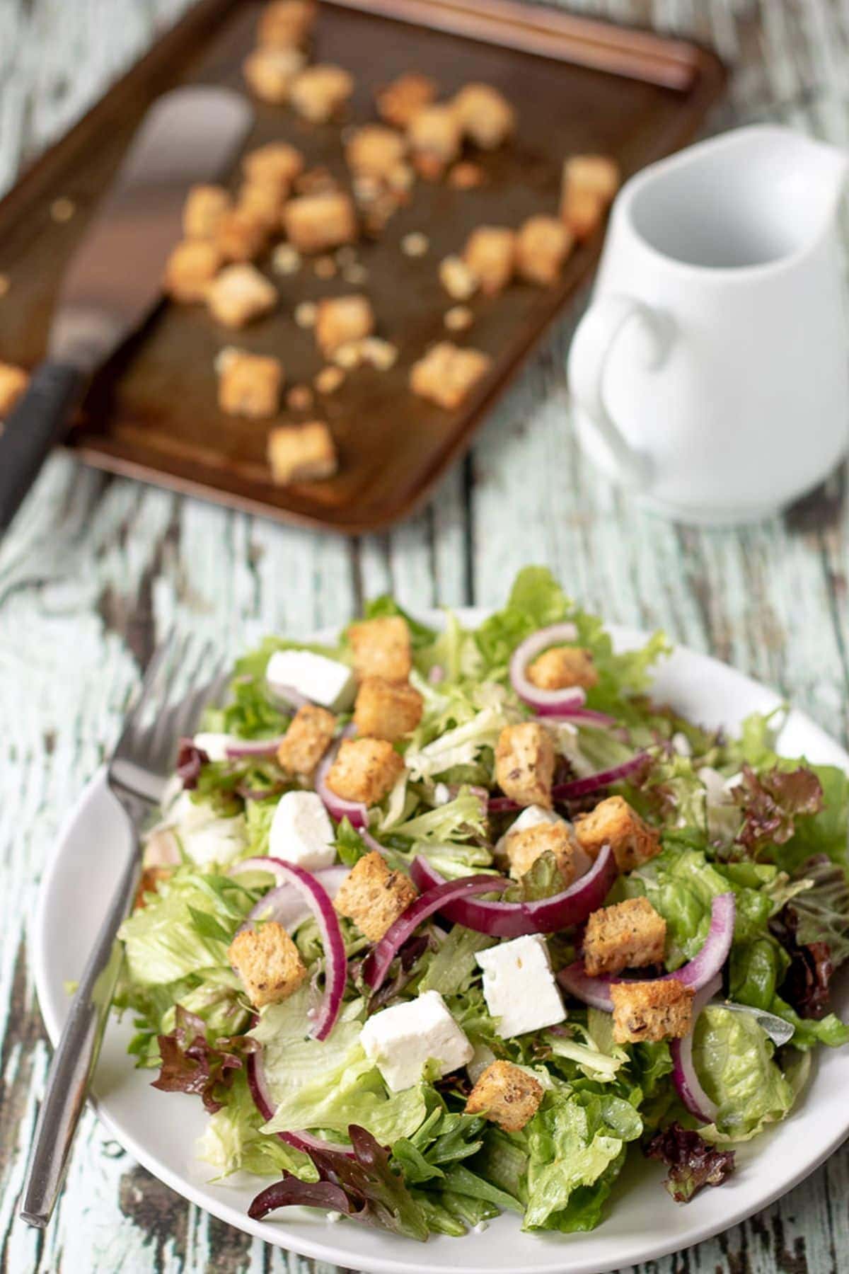 Served plate of feta and crouton salad ready to eat with a fork on the side and the tray of baked croutons in the background.
