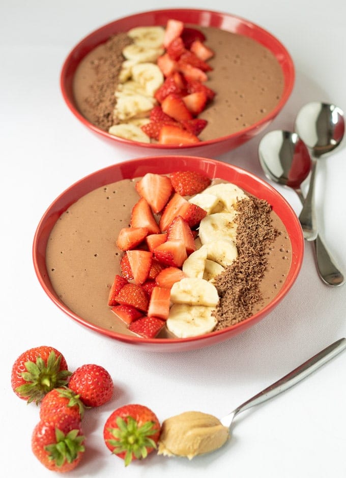 Two peanut butter chocolate banana smoothie bowls decorated with chopped strawberries, sliced bananas and grated chocolate ready to eat with two spoons. And some strawberries and a teaspoon of peanut butter in the foreground.
