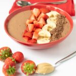 Peanut butter chocolate banana smoothie bowl decorated with chopped strawberries, sliced bananas and grated chocolate in the background with some strawberries and a teaspoon of peanut butter in the foreground.