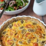 Tomato leek and mushroom quiche just taken out of the oven with bowl of size salad in the background.