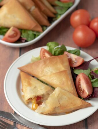 A plate of roast vegetable filo parcels and salad with one of the filo parcels cut in half with the roasted vegetable filo showing. The rest of the parcels on a serving platter in the background.