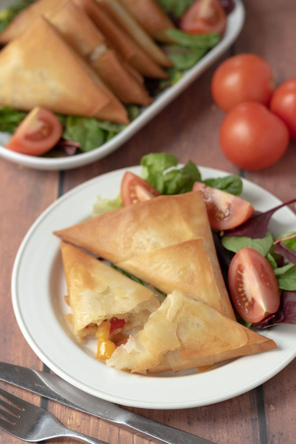 A plate of roast vegetable filo parcels and salad with one of the filo parcels cut in half with the roasted vegetable filo showing. The rest of the parcels on a serving platter in the background.