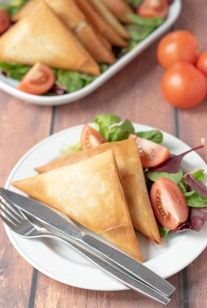 Plate of roast vegetable filo parcels with salad and a knife and fork on with a serving dish of the rest of the filo parcels in the background.