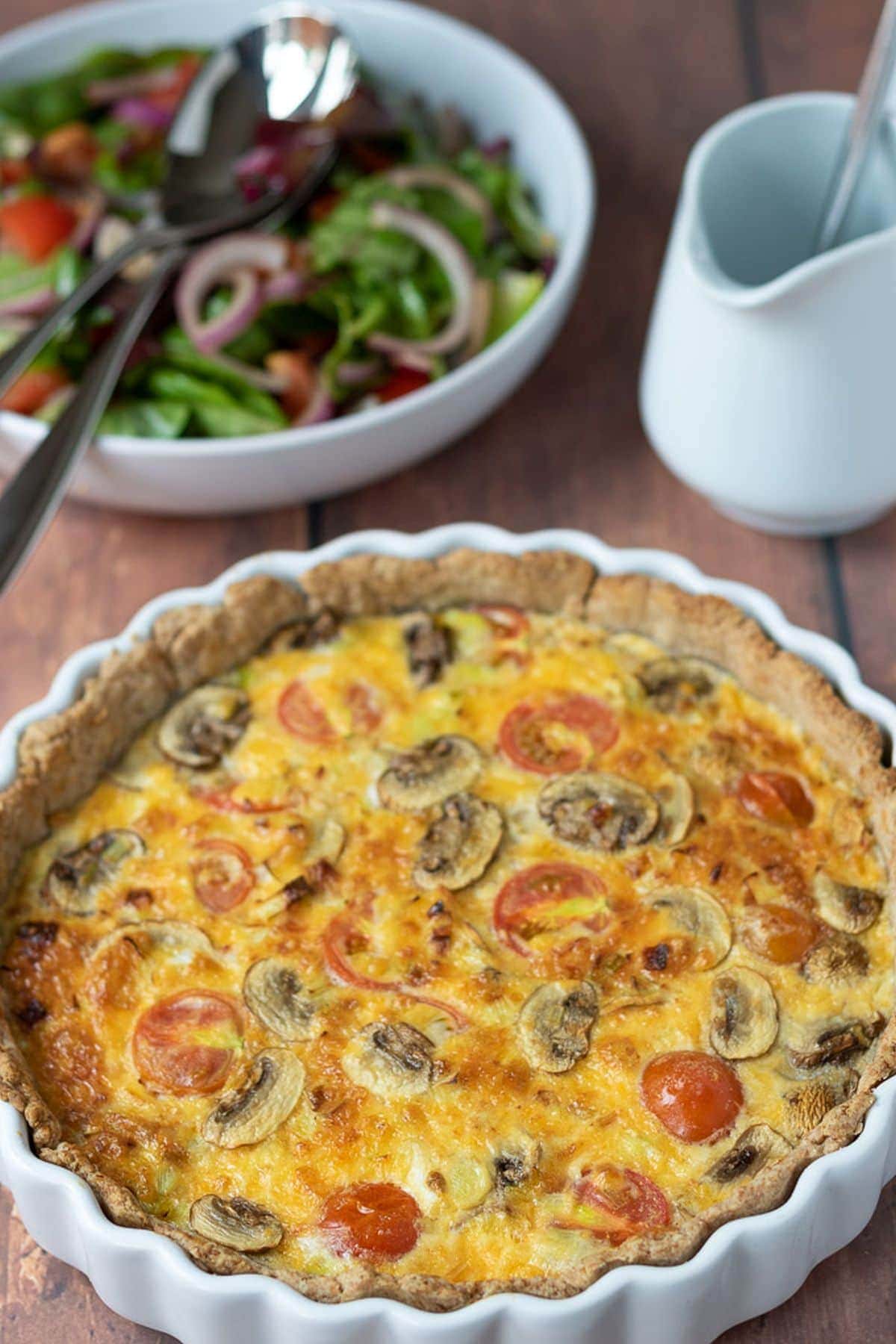 Tomato leek and mushroom quiche just taken out of the oven with a bowl of side salad and a jug in the background.