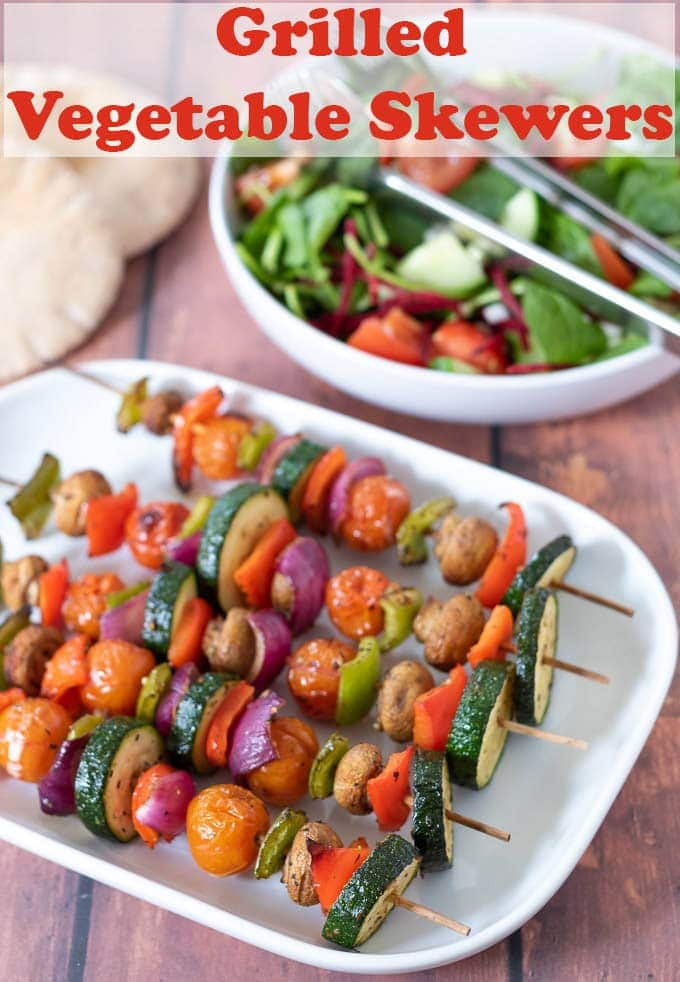 Serving dish of cooked grilled vegetable skewers with a bowl of side salad and pitta breads in the background.