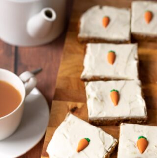 Healthy carrot cake traybake slices on a chopping board with a cup of tea and teapot to the left.