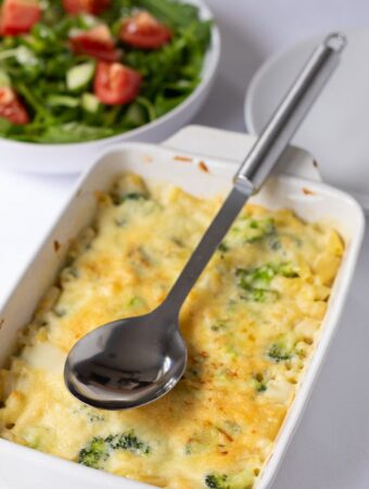 Healthy baked macaroni with broccoli casserole taken out of oven and on a table with a serving spoon on top and side salad in the background.