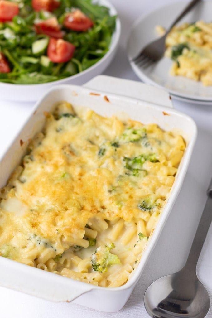 Healthy baked macaroni with broccoli on a table with a slice taken out on a seperate place and a side salad in the background.