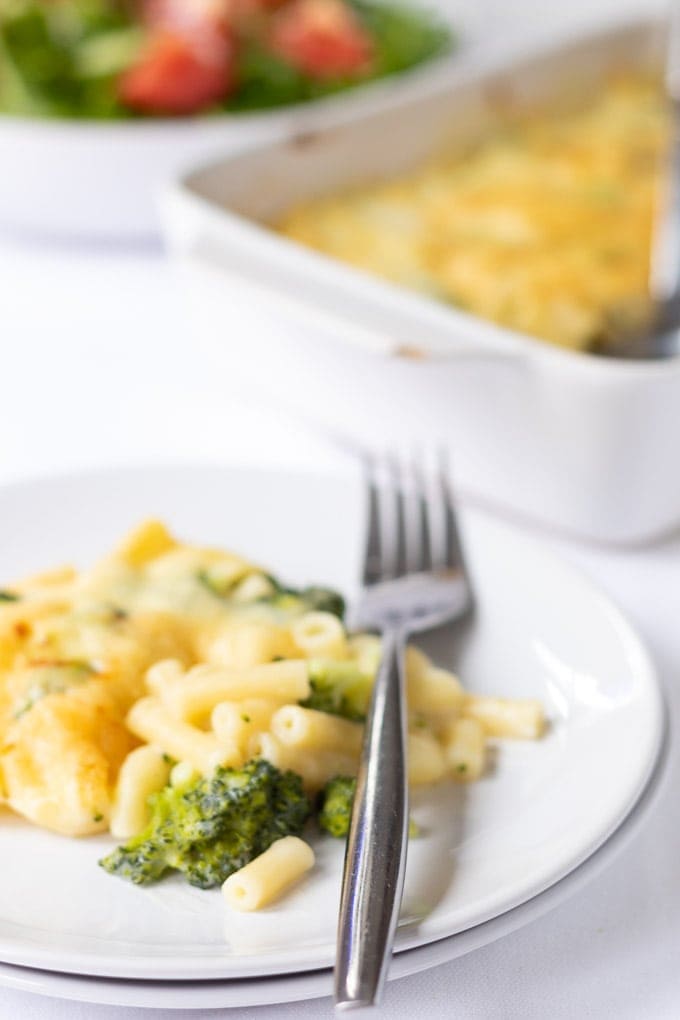 A slice of baked macaroni and broccoli on a plate. Serving dish and side salad in the background.