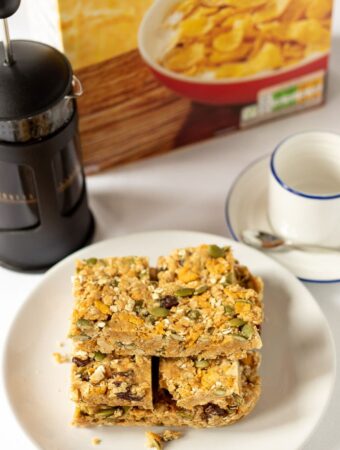 No-bake cereal breakfast bars stacked on a plate with a cafetiere, coffee cup and a box of cereal in the background.