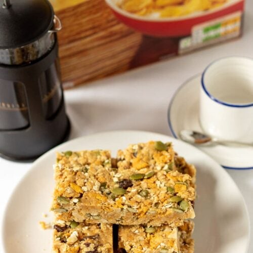 No-bake cereal breakfast bars stacked on a plate with a cafetiere, coffee cup and a box of cereal in the background.