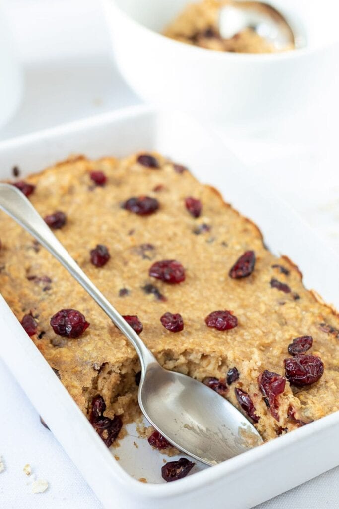 Baked cranberry oatmeal in a baking dish with a serving taken out and a serving spoon in the dish.