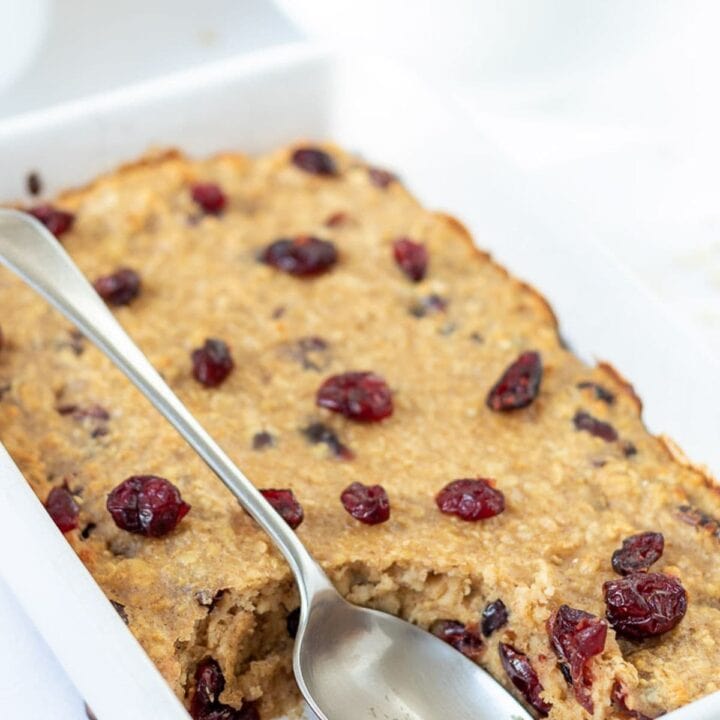 Baked cranberry oatmeal in a baking dish with a serving taken out and a serving spoon in the dish.