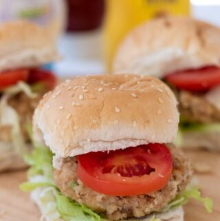 Three leftover chicken burgers in buns with iceberg lettuce and sliced tomatoes. All sitting on a bread board with condiments in the background.