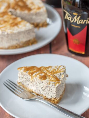 A slice of no-bake Tia Maria cheesecake on a plate with the full cake and a bottle of Tia Maria in the background.