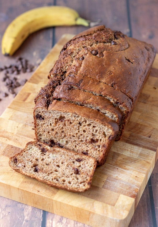 Chocolate chip banana loaf cake on a bread board with 4 slices cut off. A banana and chocolate chip decorations at the back.