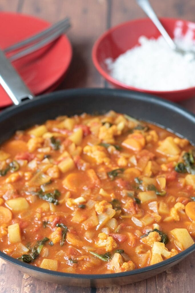 A pan of cooked easy mixed vegetable curry with a dish of rice and serving plates in the background.