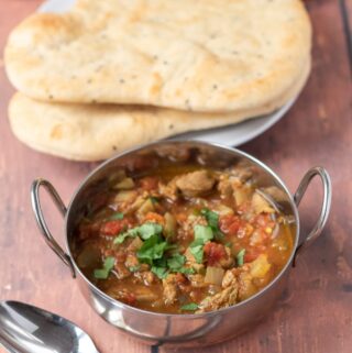 A serving of slow cooker lamb curry in a balti dish with 2 naan breads in the background.