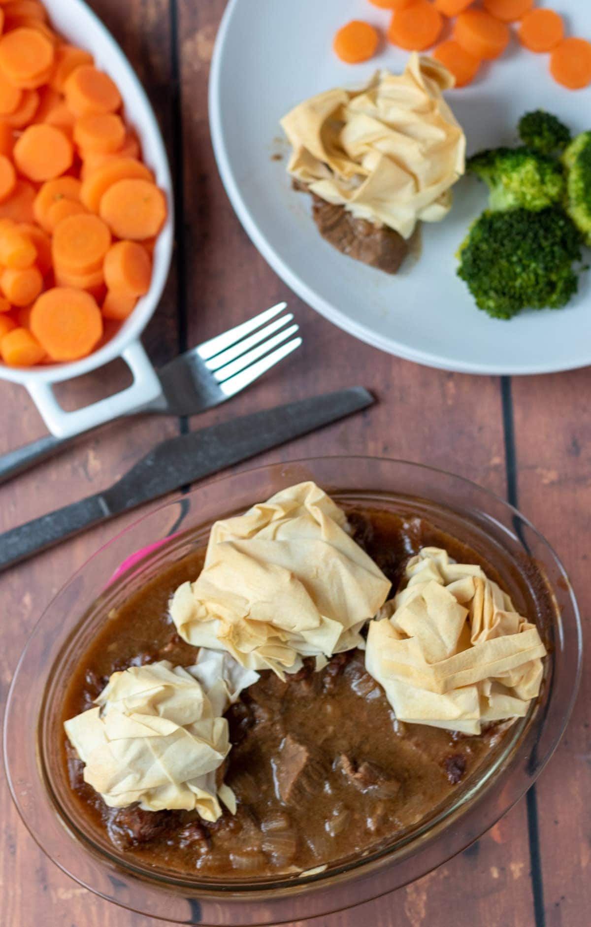 Bired eye view of steak pie with filo pastry on top. A portion has been removed and plated in the background with broccoli and carrot sides.