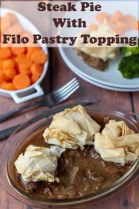 Steak pie with filo pastry topping and sides of broccoli and carrots in serving dishes in the background. Pin title text overlay at top.