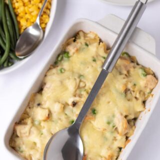 Birds eye view of a cooked casserole dish of honey mustard chicken pasta bake with a serving spoon on top. With green beans and sweetcorn in a side dish.