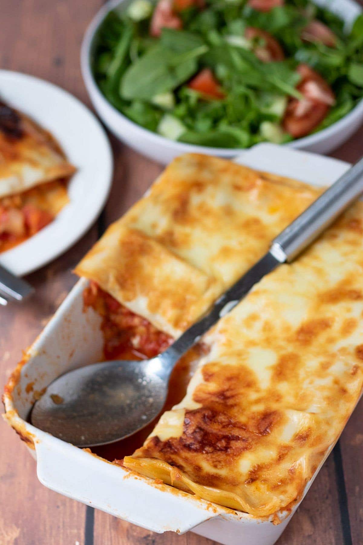 Red lentil lasagne with a portion taken out of one corner. A spoon in its place and a side salad in the background.