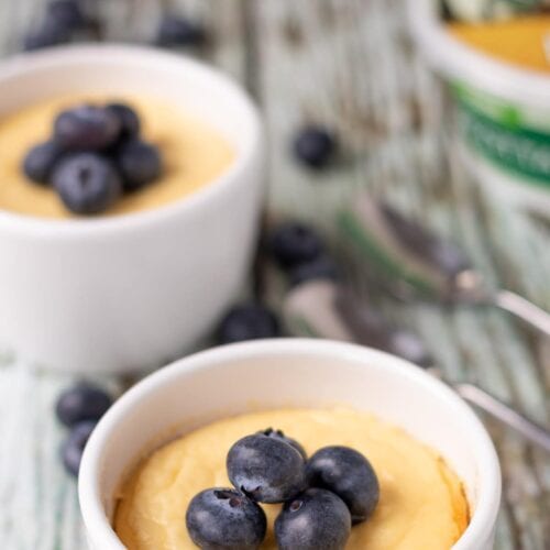 Two baked vanilla ricotta pudding ramekins diagonally on front of each other topped with blueberries.