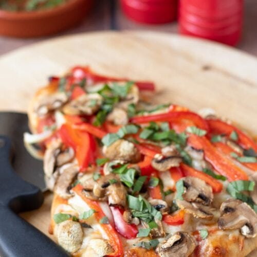 Vegetarian naan pizza cooked and placed on a round chopping board with a pizza cutter to the side. Salt and pepper and basil to garnish in the background.