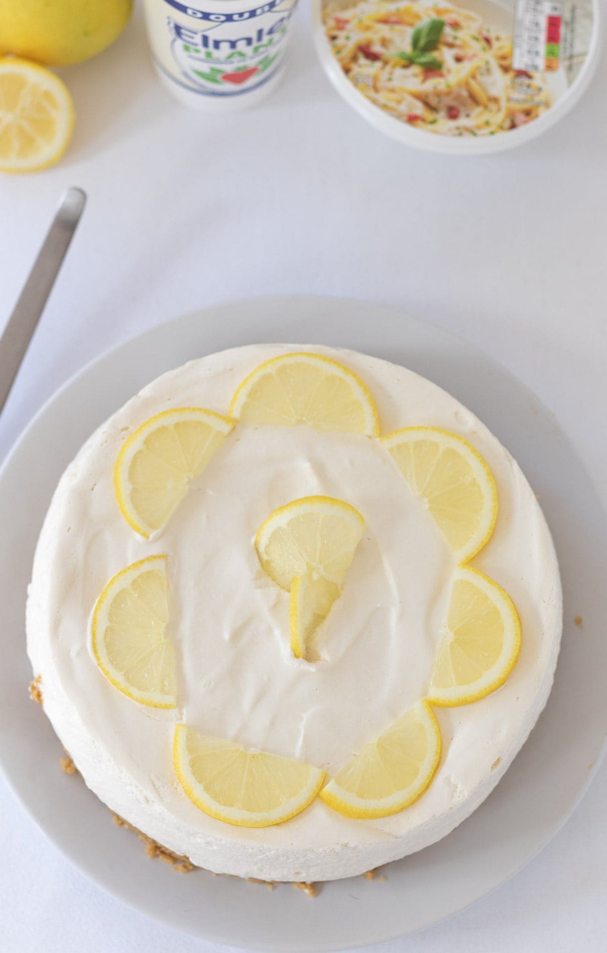 Birds eye view of a dairy free cheesecake decorated with slices of lemon with alternative dairy free product containers and a sliced lemon in the background.