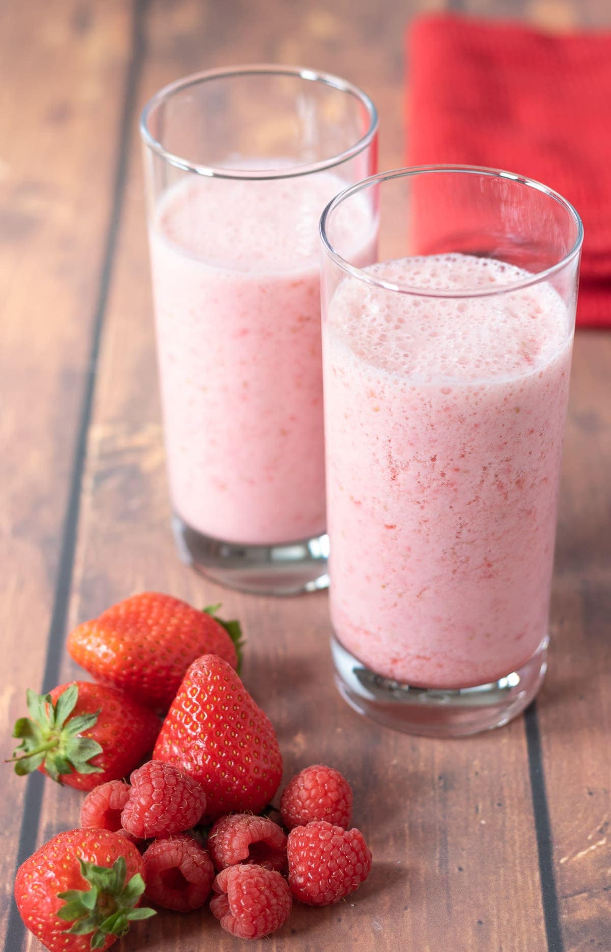 Two tall glasses of strawberry and raspberry smoothie in the center with a pile of strawberries and raspberries in front of the glasses. Serviette to the rear.