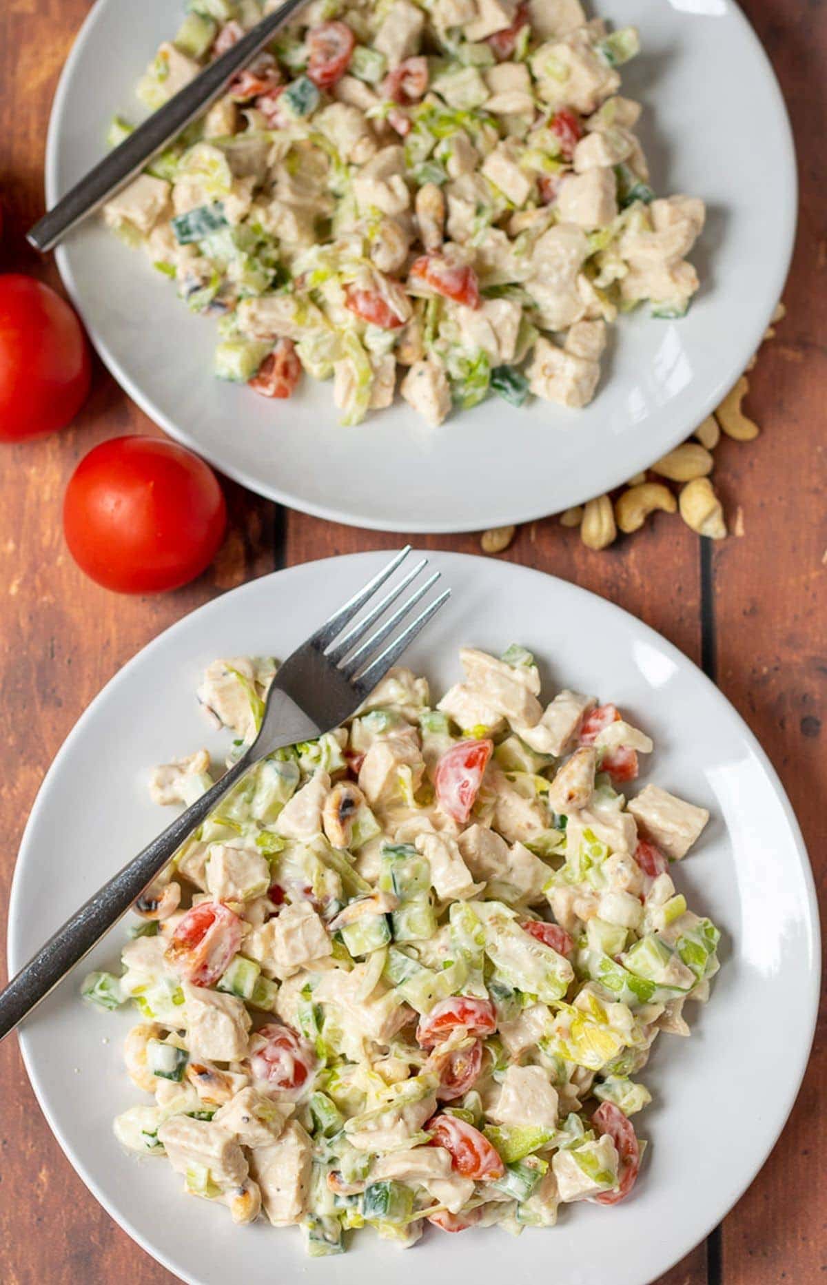 Birds eye view of two plates of healthy chicken salad with forks on. 2 tomatoes and cashew nuts in between as decoration.