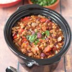 Slow cooker vegetarian chilli in slow cooker. Dishes of rice and coriander at the top.