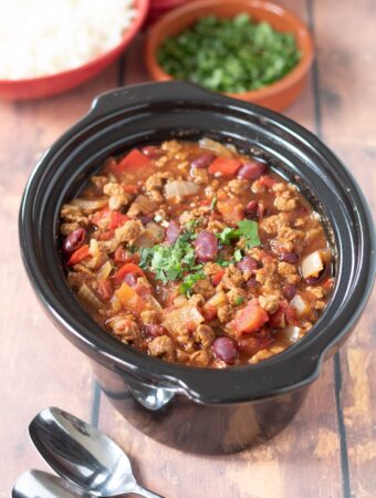 Slow cooker vegetarian chilli in slow cooker. Dishes of rice and coriander at the top.